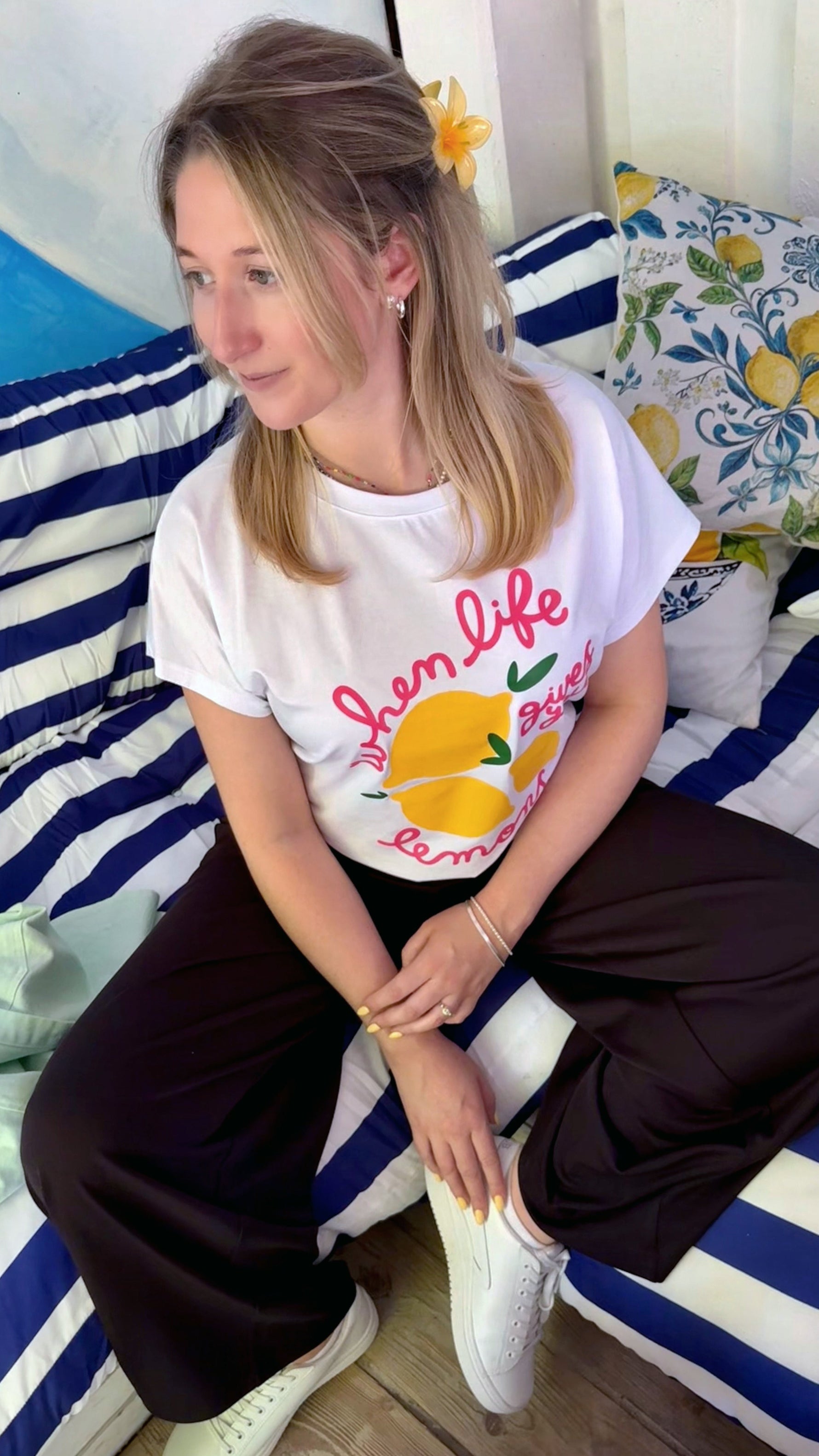 Fashion Editorial in italienischem Flair: junges Model trägt ein ausgefallenes T-Shirt mit pinker Schrift und gelben Zitronen. Sie sitzt im Urlaub im Strandcafé auf einem Sofa mit Blau Weißen Streifen.