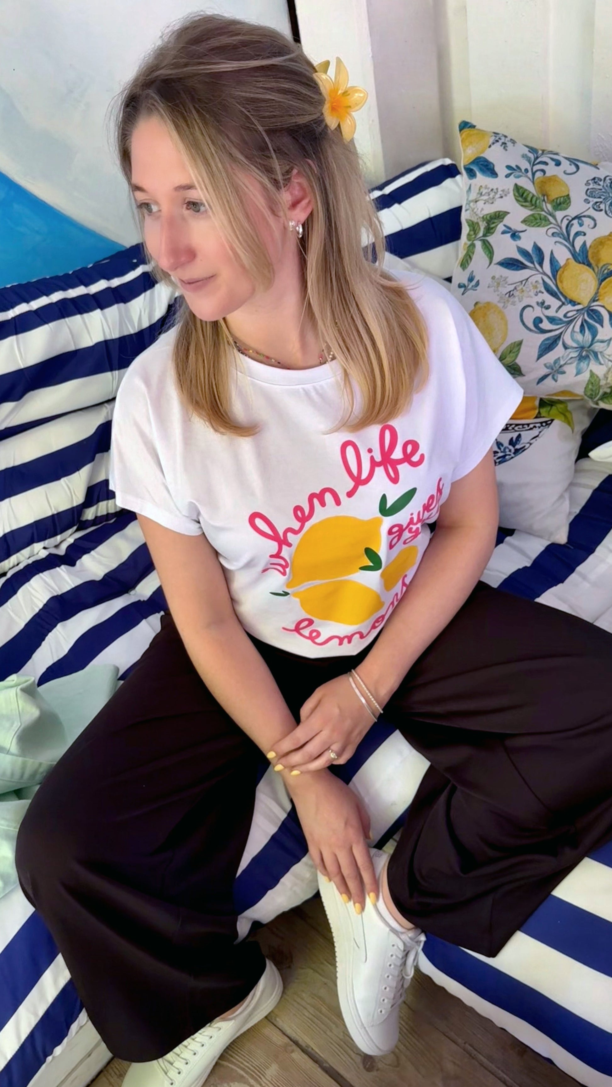 Fashion Editorial in italienischem Flair: junges Model trägt ein ausgefallenes T-Shirt mit pinker Schrift und gelben Zitronen. Sie sitzt im Urlaub im Strandcafé auf einem Sofa mit Blau Weißen Streifen.