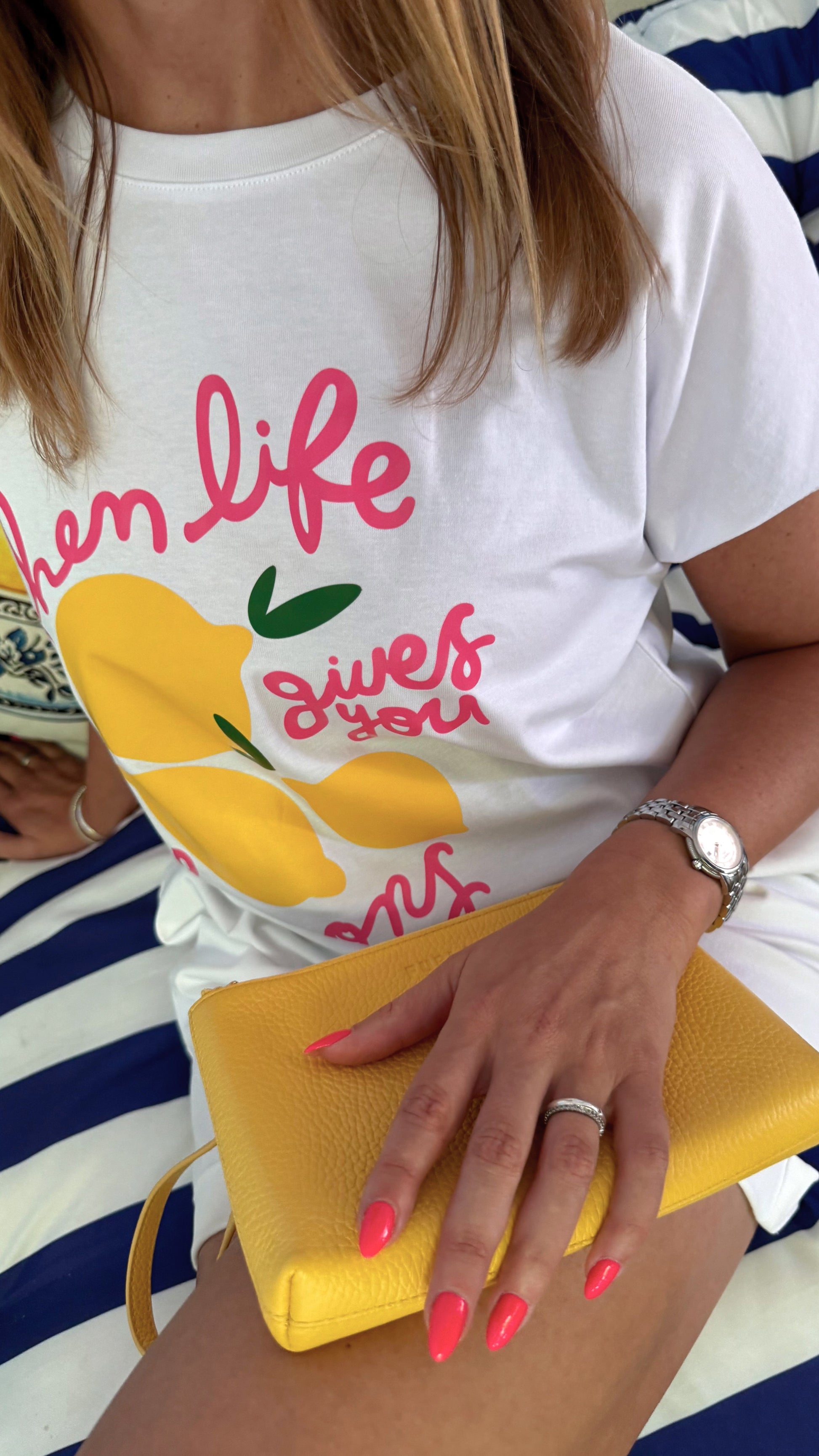 Fashion Editorial in italienischem Flair Pink-Gelb- Blau: Detailzoom auf T-Shirt  : Model trägt ein ausgefallenes T-Shirt mit pinker Schrift und gelben Zitronen. Sie sitzt im Urlaub im Strandcafé auf einem Sofa mit Blau Weißen Streifen.