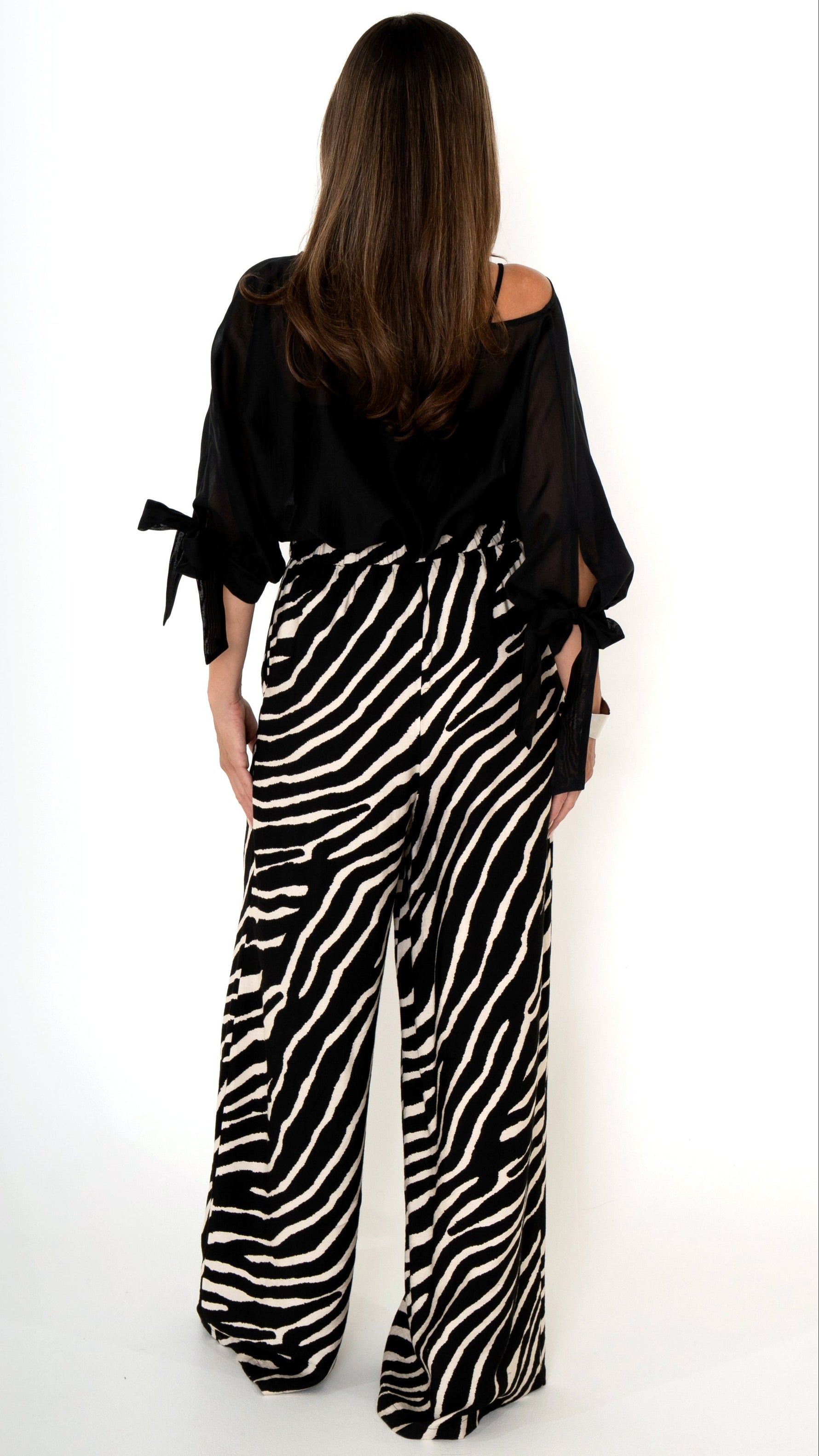 Rückansicht einer eleganten ausgefallenen Damenbekleidung in schwarz weiß: Modisch sommerliches Outfit bestehend aus einer weiten Zebra Print Hose und einer schwarzen Chiffonbluse  mit Schleifen am Ärmel, Halbarm