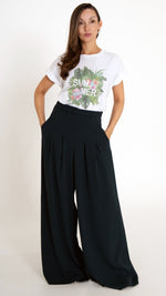 Feminine ausgefallene Damenmode: Model trägt ein weißes Sommer T-Shirt mit tropischem Palmendruck. Dazu trägt sie eine modische Stoffhose mit ultra weitem Bein in Petrol.