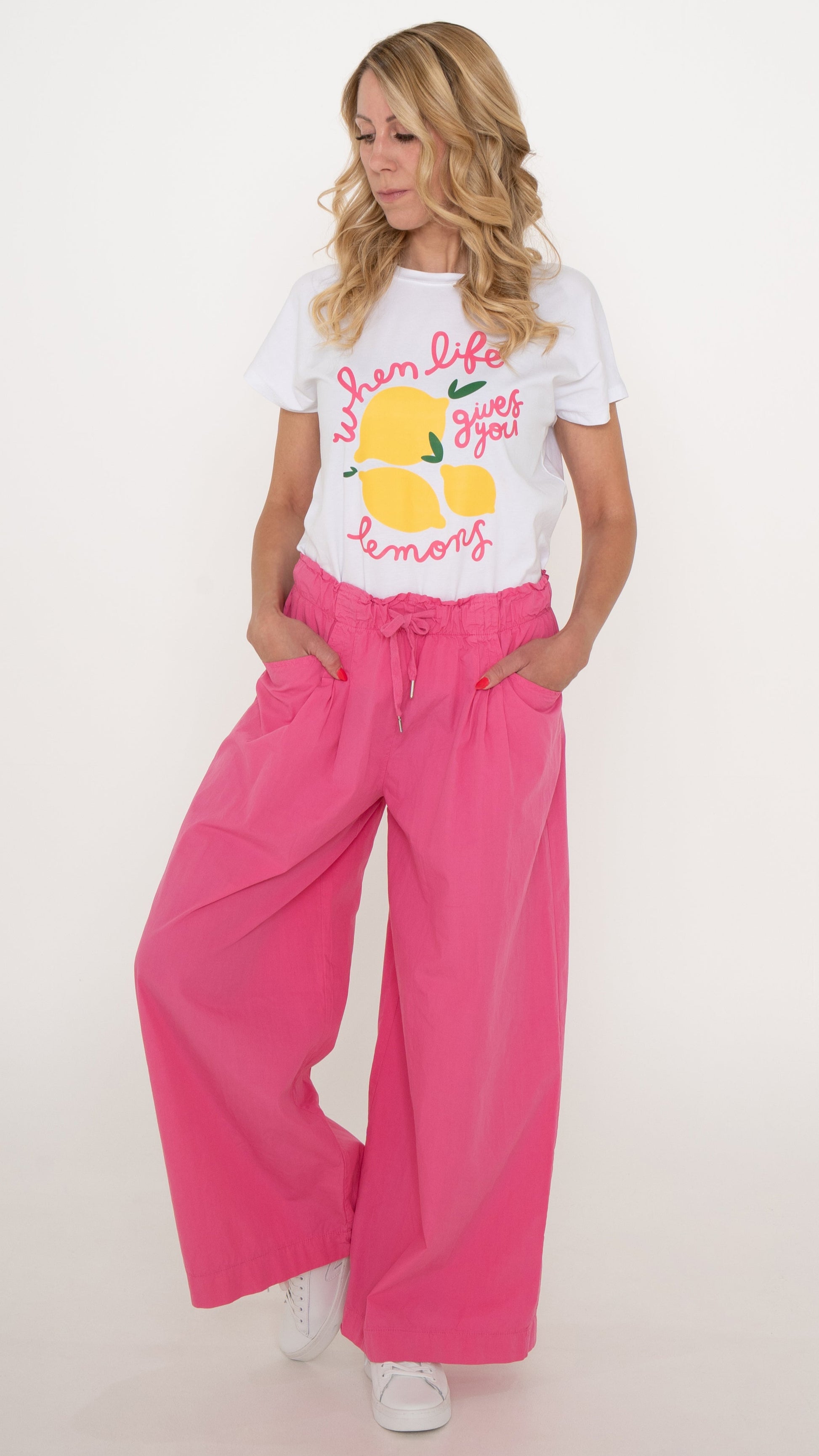 Feminine coole Damenmode: Das Model trägt ein cooles ausgefallenes T-Shirt in weiß mit pinker Schrift und gelben Zitronen. Dazu trägt das Model eine sommerlich lässige Stoffhose in knalligem Pink mit Falten und weitem Bein. Bindeband