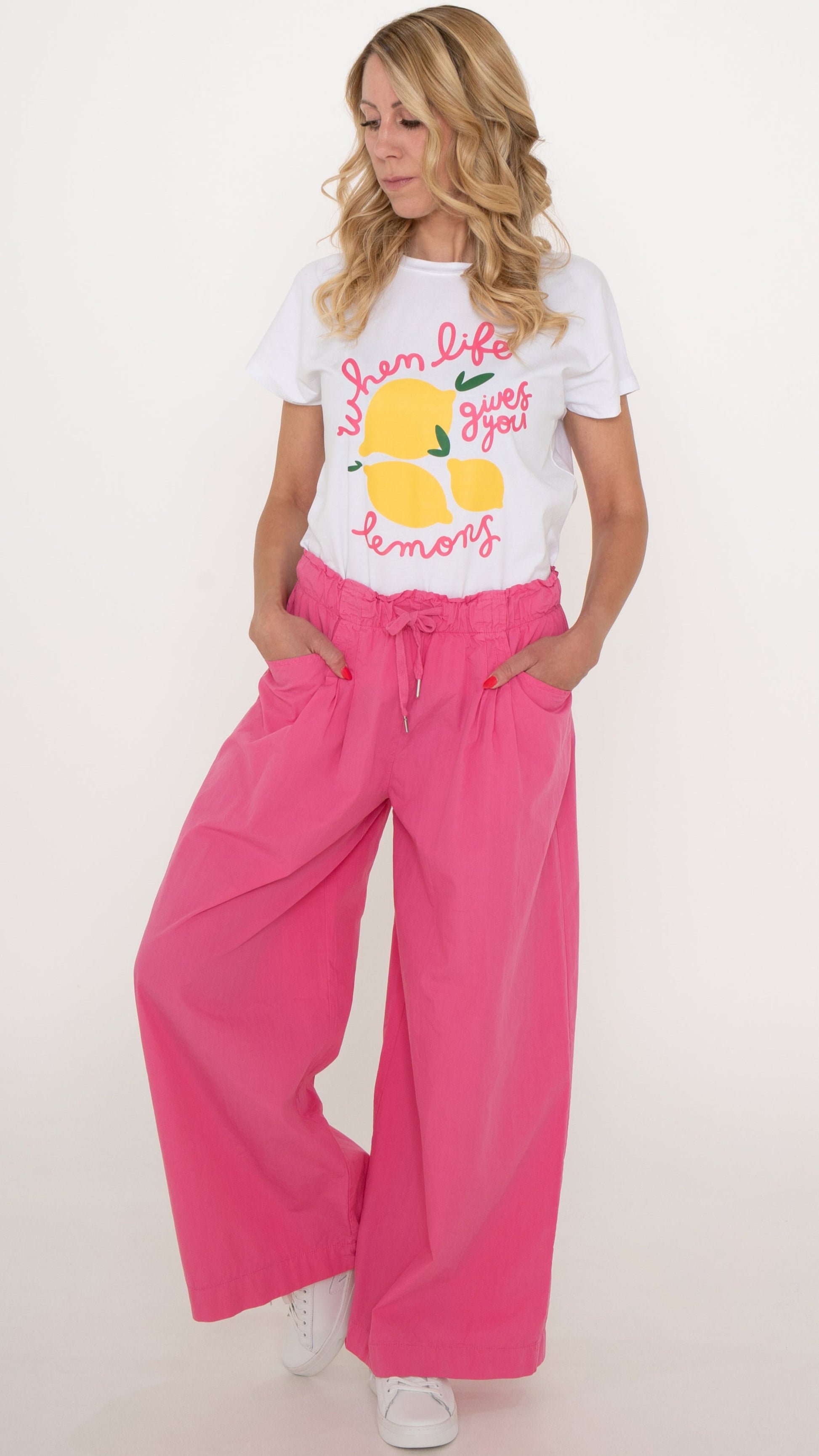 Eine blonde Frau trägt ein cooles lässiges Sommeroutfit: pinke weite Damen Hose mit elastischem Bund und Schleife, lässige Passform. Dazu ein weißes Sommer T-shirt mit Druck aus Zitronen und pinker Schrift