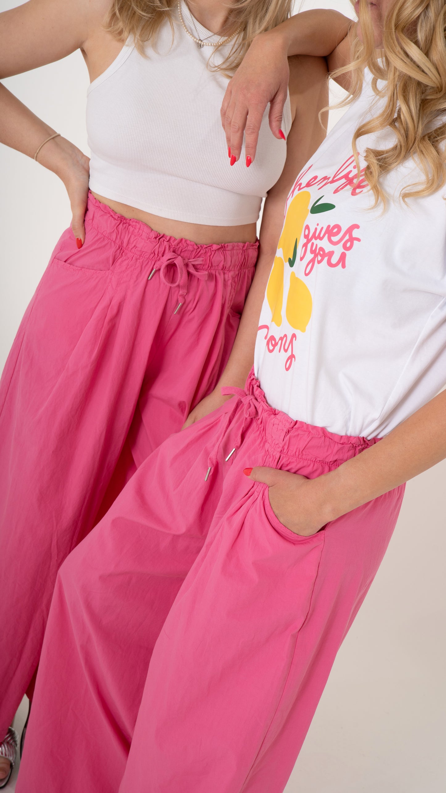 2 Damen tragen ein lässig cooles Sommeroutfit: Fokus des Bildes ist die pinke lässig weite Hose mit Eingriffstasche und elastischem Bund. Dazu ein weißes Oberteil