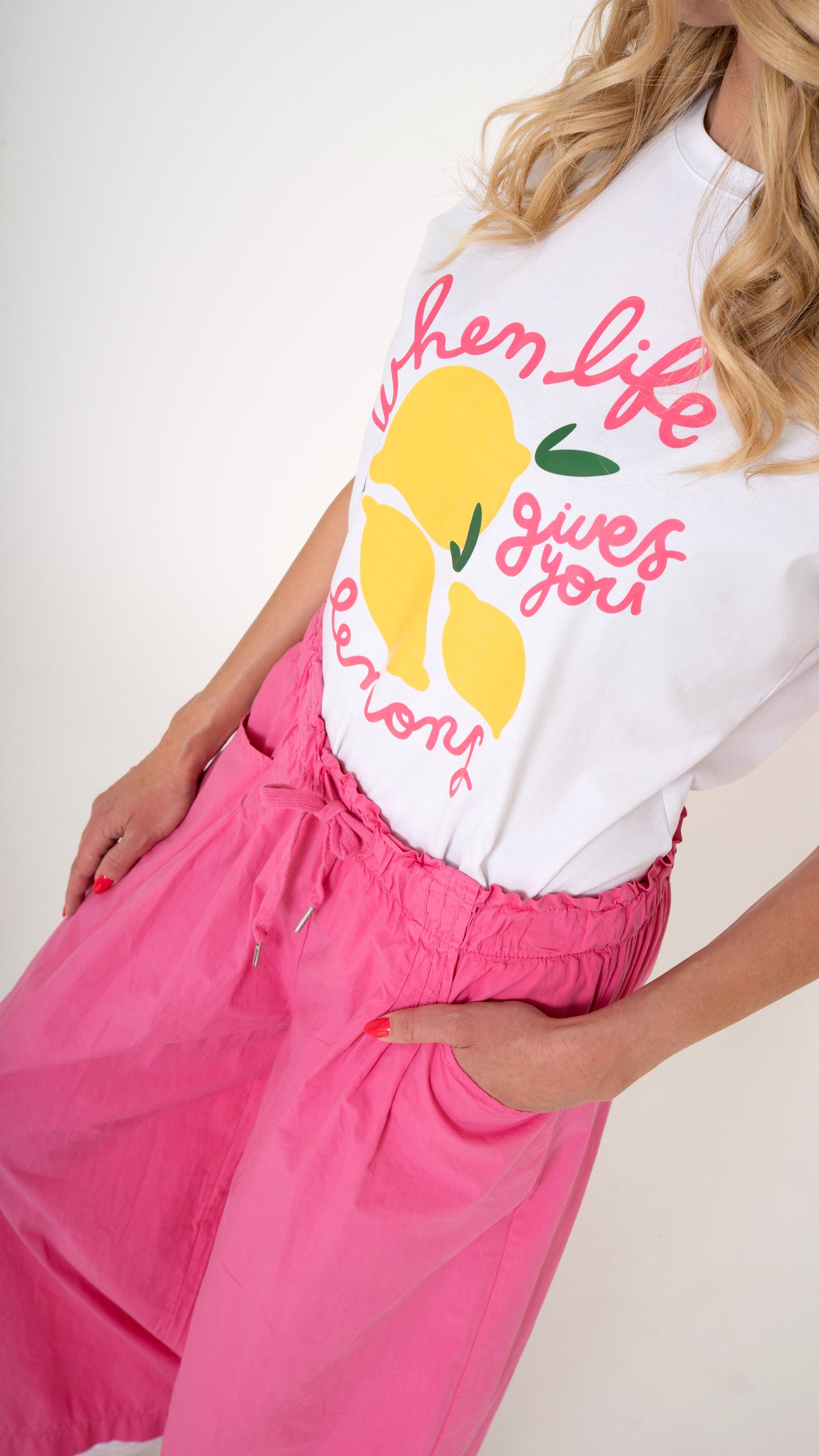 Eine blonde Frau trägt ein cooles lässiges Sommeroutfit: pinke weite Damen Hose mit elastischem Bund und Schleife, lässige Passform. Dazu ein weißes Sommer T-shirt mit Druck aus Zitronen und pinker Schrift