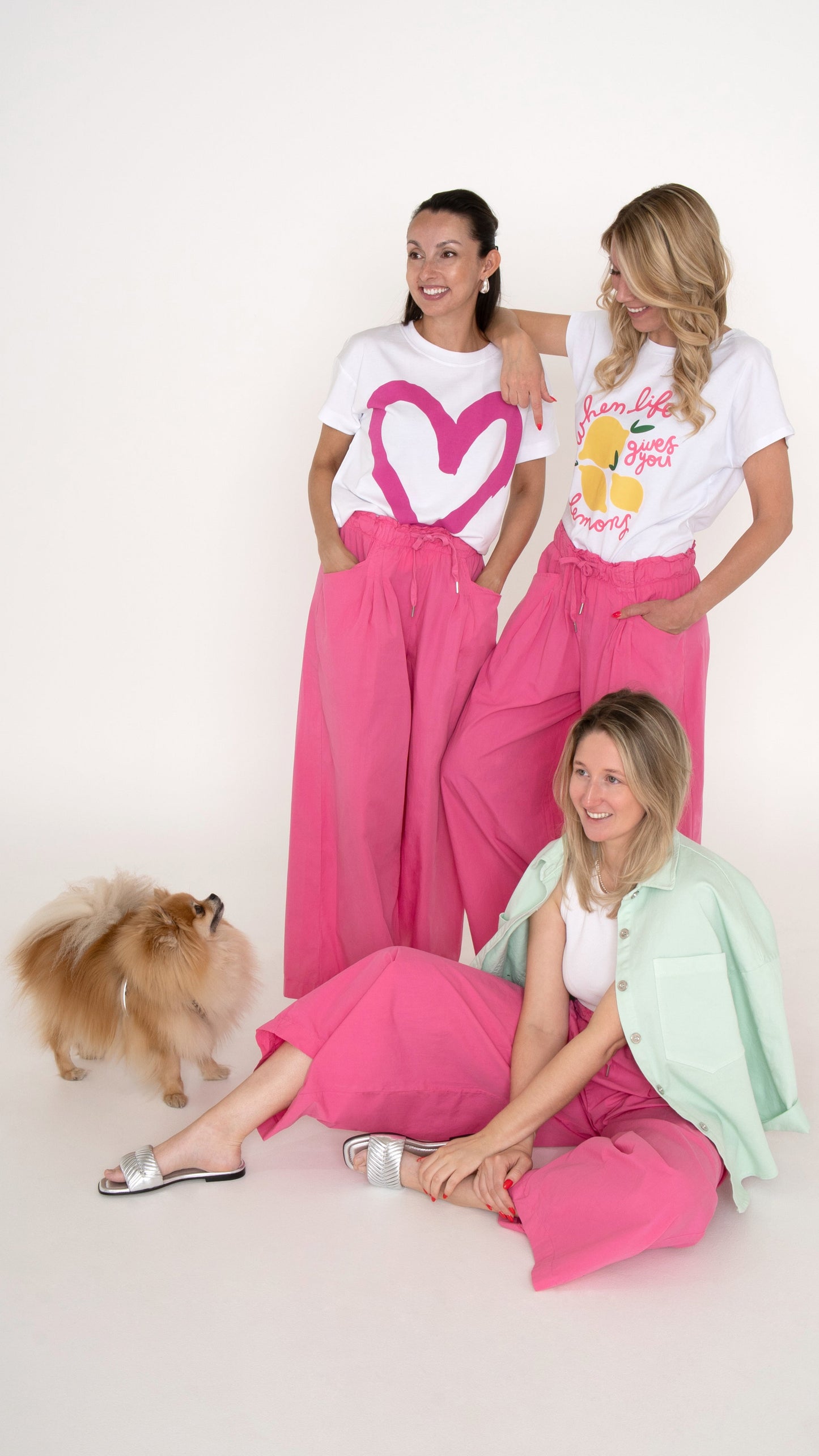3 Freundinnen tragen ein sommerlich ausgefallene Damenkleidung mit einer weiten pinken Hose. Dazu tragen sie ein weißes Oberteil mit und ohne Print. Sie sind fröhlich und lachen. Ein kleiner Hund begleitet sie