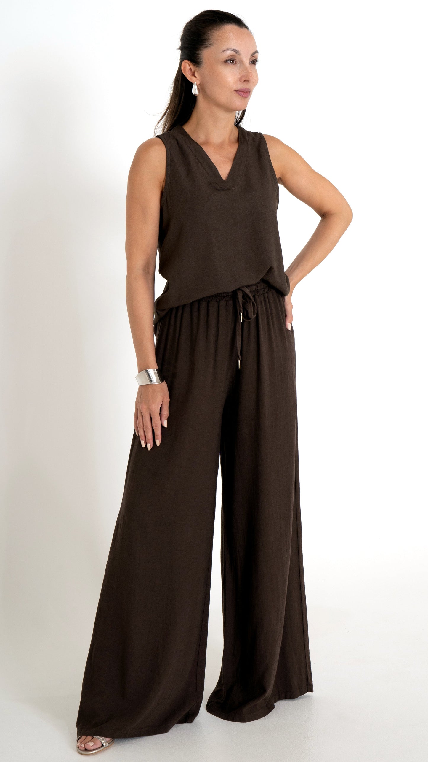 Eine Dame mit dunklen Haaren trägt ein sommerlich schickes Outfit in der Farbe Mocca braun. Es besteht aus einer weiten lockeren Leinenhose mit Bindeband und elastischem BUnd. Das Oberteil besteht aus einem Leinentop mit V-Ausschnitt ohne Ärmel.