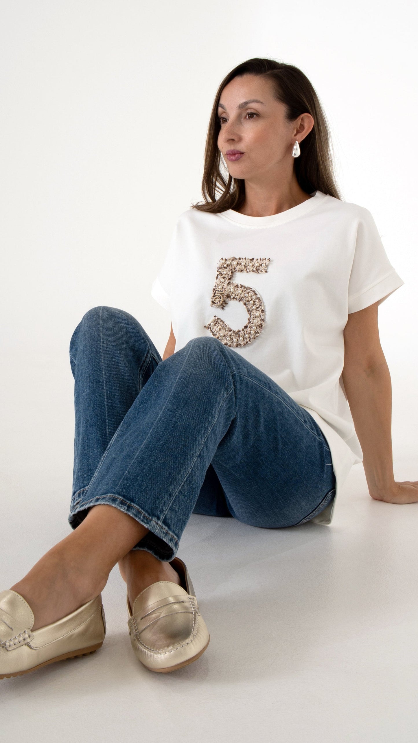 Feminine Damenmode sitzende Ansicht: Model trägt ein feminines Alltagsoutfit bestehend aus einer geraden Denim Jeanshose in blau, goldene metallic Loafer und ein modernes T-Shirt mit lockerer Passform und einer Stickerei in Form einer 5