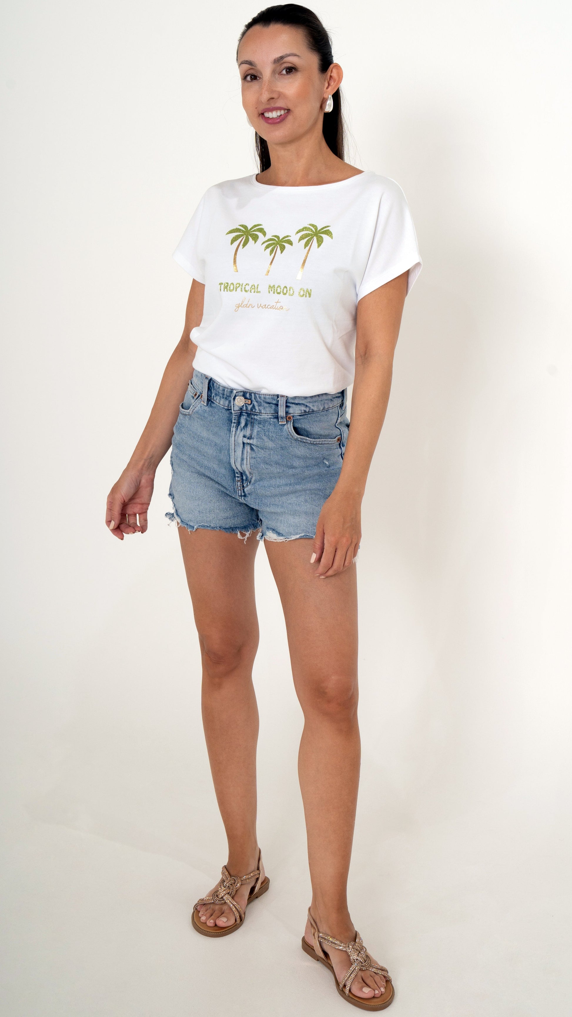 Sommerliche  feminine Damenmode: Das Model trägt ein sommerliches Outfit bestehend aus einer blauen Jeans Short und ein weißes T-Shirt mit tropischem Palmenprint mit Gold Glitzer Sandalen